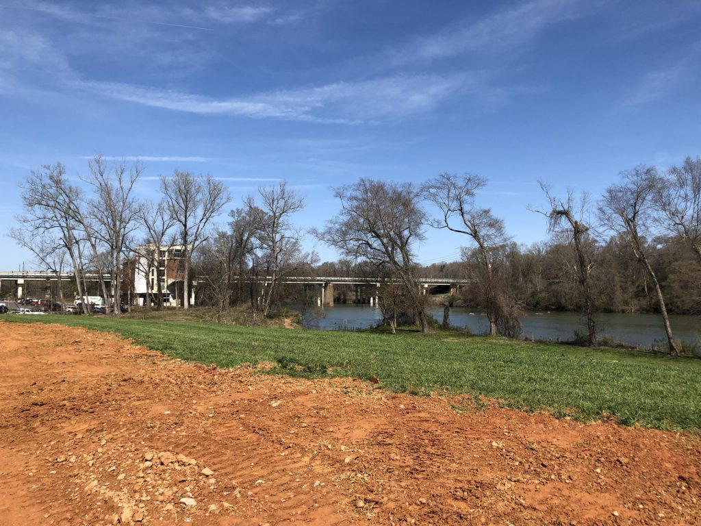 A sweeping vista of the Catawba River opens up along the Carolina Thread Trail, with a bridge and historic mill silhouetted beneath wispy clouds.
