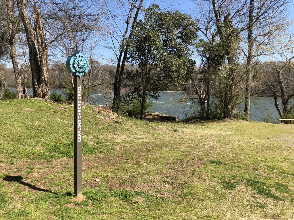A Carolina Thread Trail marker stands sentinel on the grassy bank of the Catawba River, where the Riverwalk connects Rock Hill to a wider regional trail network.