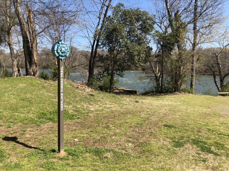 A Carolina Thread Trail marker stands sentinel on the grassy bank of the Catawba River, where the Riverwalk connects Rock Hill to a wider regional trail network.