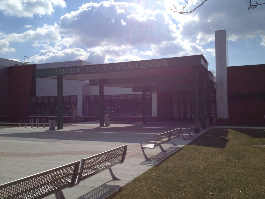 Sunlight streaming over the Farmingdale Campus Center entrance with empty benches in the foreground and a tower sign on the right.
