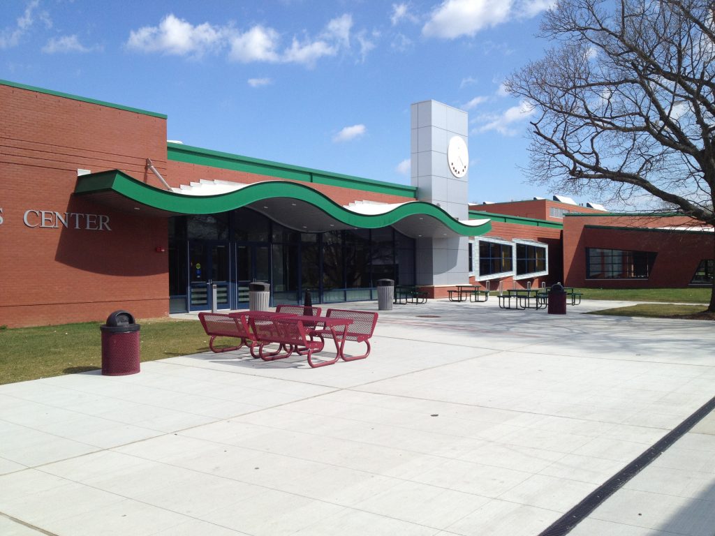 Red picnic tables on the Campus Center plaza beneath a green wave canopy, with a clock tower rising above the building.