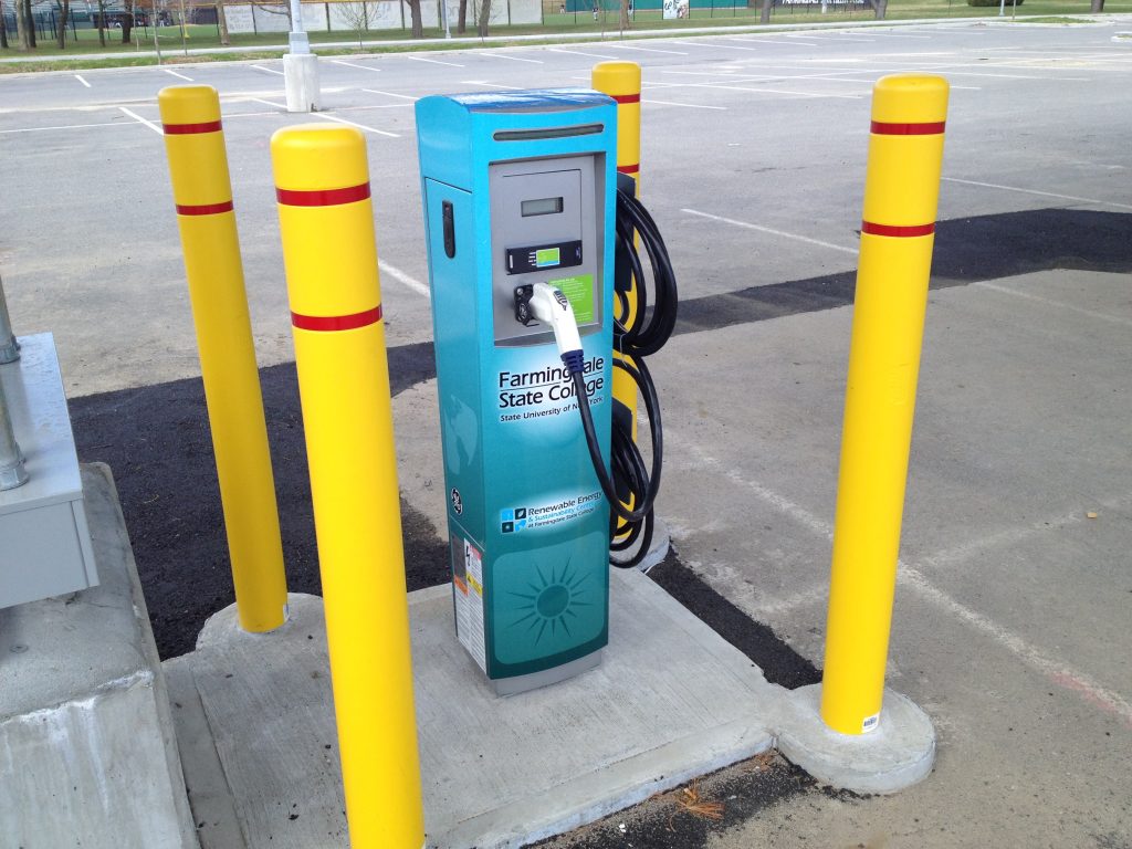A teal Farmingdale State College EV charging station with a sun graphic, ringed by yellow safety bollards in a parking lot.