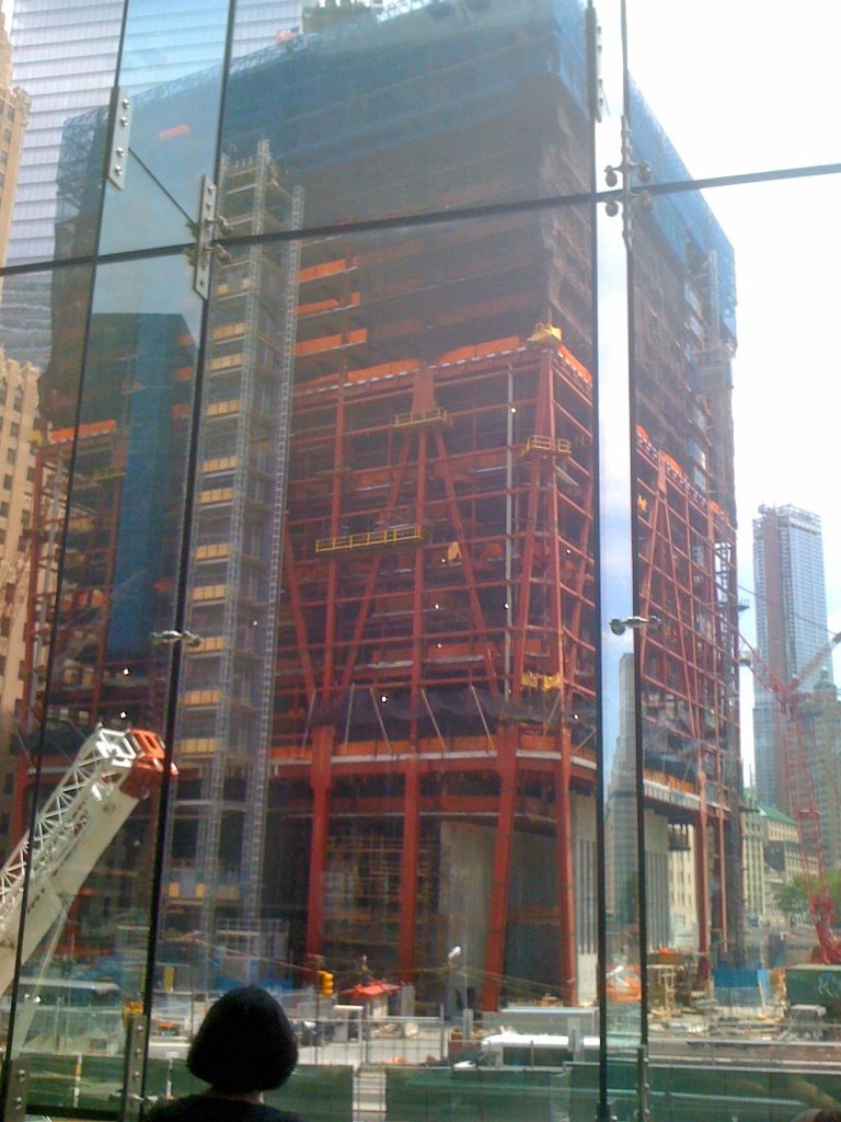 The lower floors of One World Trade Center encased in vivid orange steel scaffolding, photographed through the glass panels of the WTC viewing wall.