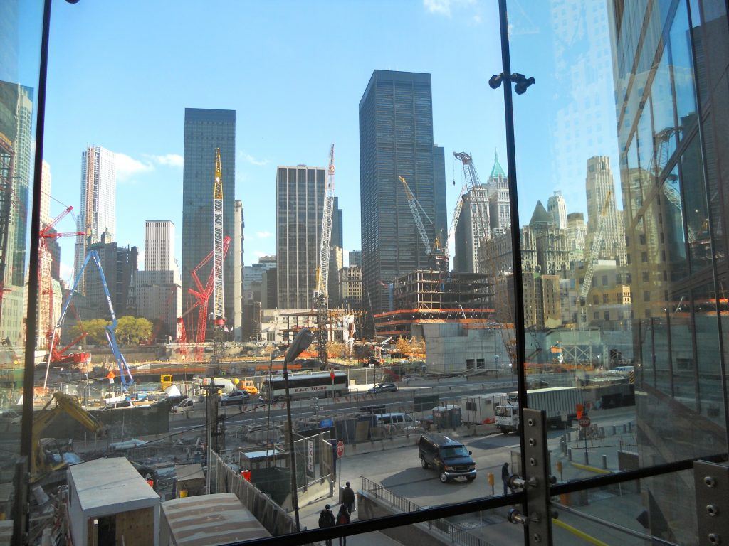 View of the World Trade Center construction site through a glass viewing wall, showing multiple cranes and the Lower Manhattan skyline
