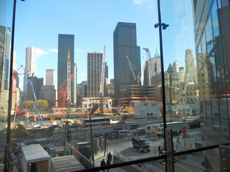 The World Trade Center construction site, photographed through a glass viewing panel, reveals the scale of the rebuilding effort with multiple cranes against the Lower Manhattan skyline.