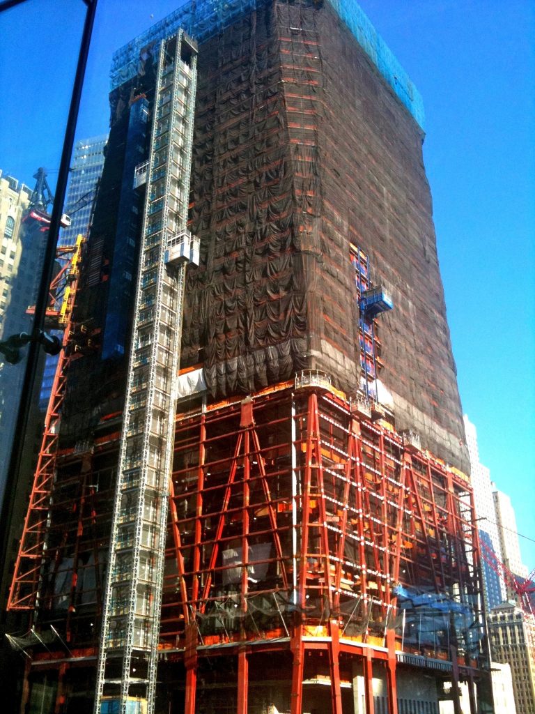 Close-up of One World Trade Center during construction, the tower wrapped in dark safety netting and braced by vivid orange steel scaffolding.