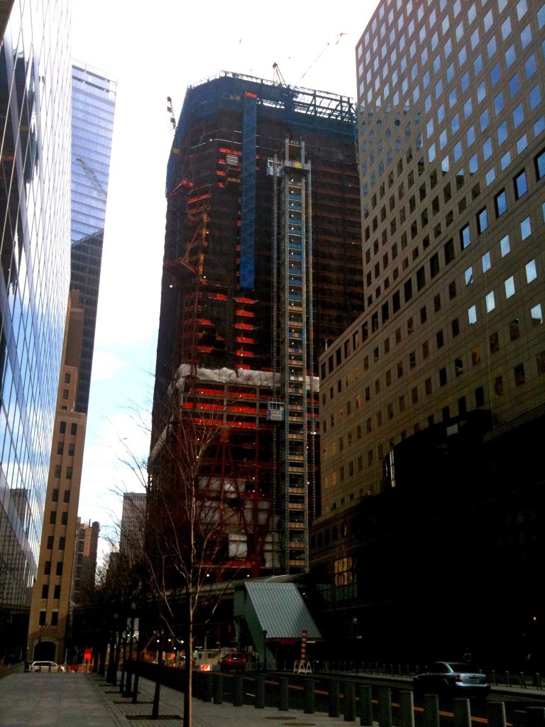 One World Trade Center tower under construction with orange scaffolding, framed between neighboring glass skyscrapers