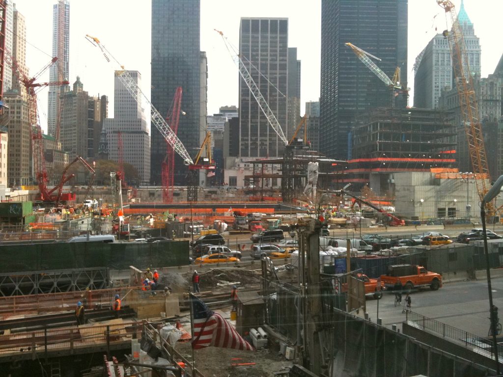 Aerial view of the World Trade Center construction site with multiple cranes, an American flag, and surrounding Manhattan skyscrapers