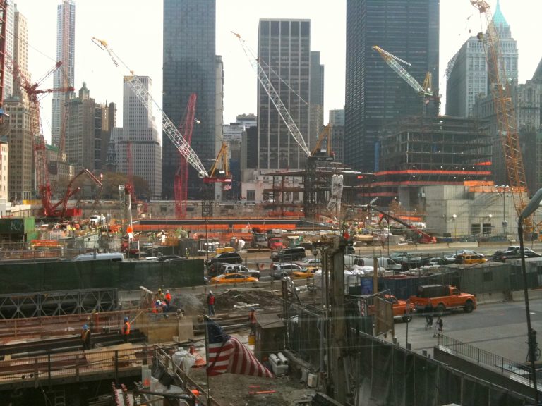 A panoramic view of the World Trade Center construction site, with tower cranes filling the air and an American flag flying at the heart of the rebuilding effort.