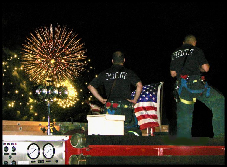 Two FDNY firefighters stand atop their fire truck, watching the Macy's 4th of July fireworks burst over the Hudson River while an American flag drapes behind them.