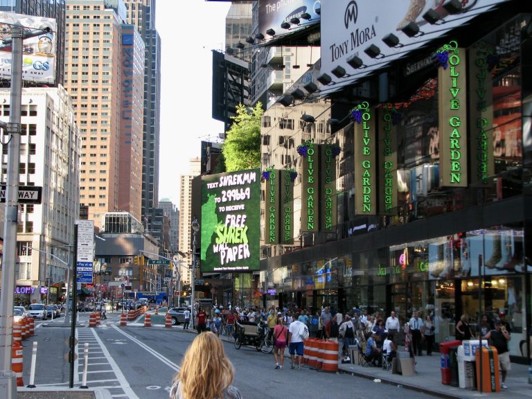 Looking down 7th Avenue in Midtown Manhattan, giant Olive Garden and Shrek the Musical billboards line the street on a busy summer day, 2009.