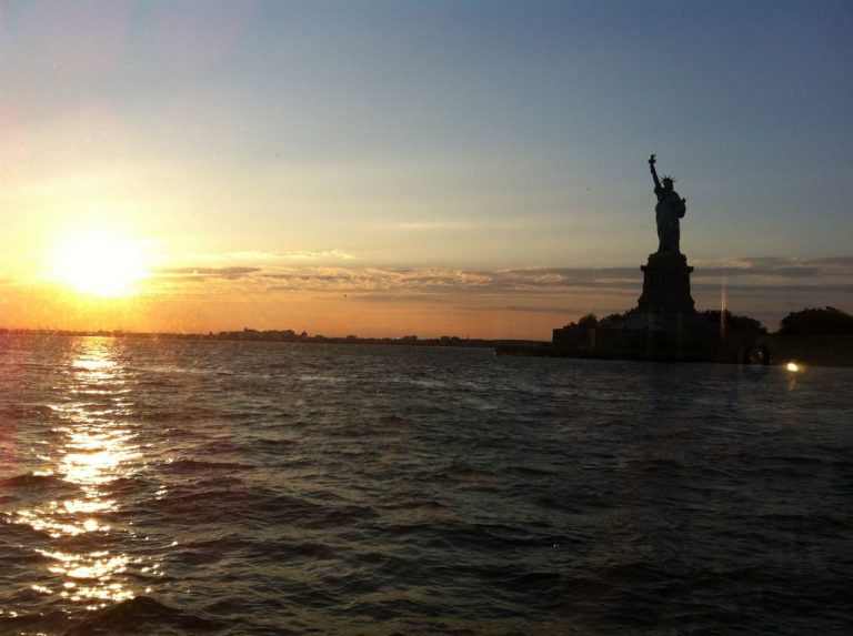 The Statue of Liberty silhouetted against a glowing sunset over the harbor.