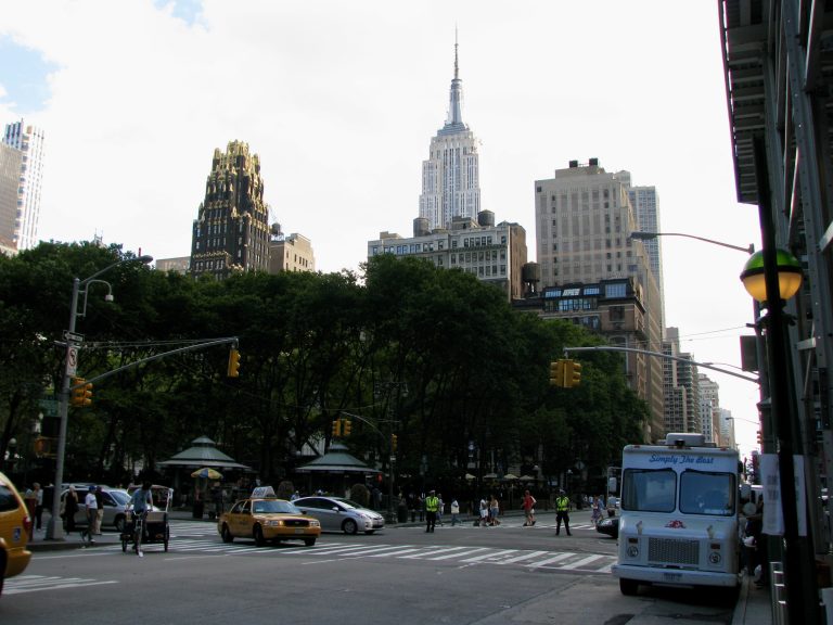 The Empire State Building towers over Midtown Manhattan, visible from a street-level view with yellow cabs and a "Simply the Best" truck in the foreground, New York City, July 4th weekend, 2009.