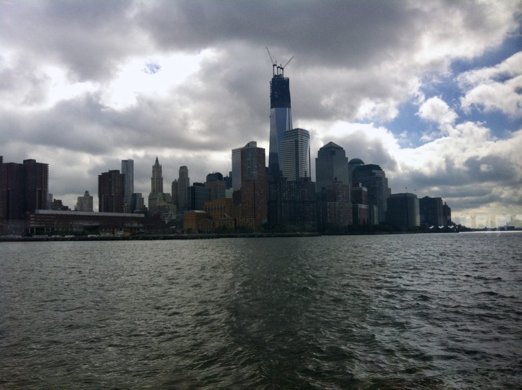 Lower Manhattan skyline viewed from the Hudson River, with One World Trade Center under construction beneath a dramatic cloudy sky