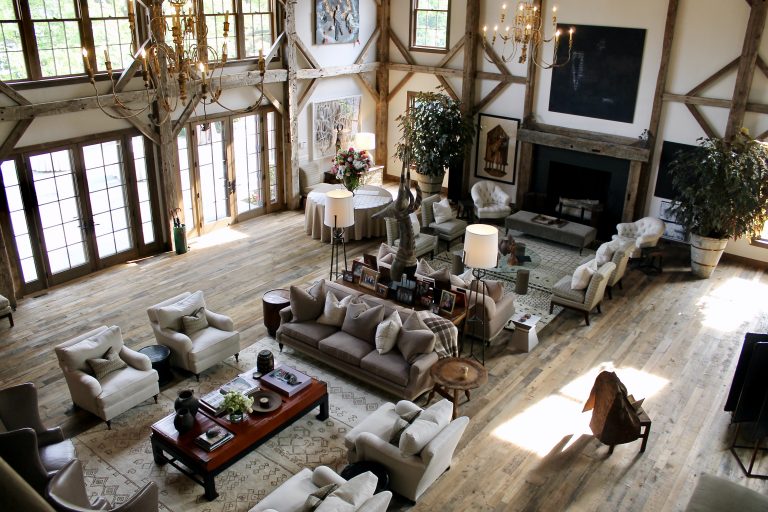 View of the great room as seen from the upstairs den, showcasing the home's double-height windows, twin chandeliers, stone fireplace, and curated seating areas.