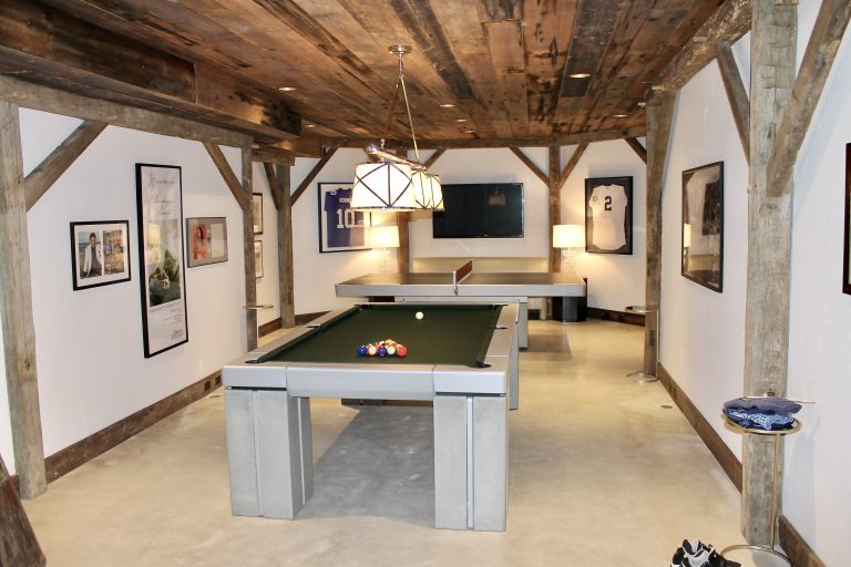 A rustic-modern game room featuring a full-size pool table and ping-pong table, framed sports jerseys, and personal memorabilia, set beneath an exposed timber-beam ceiling.