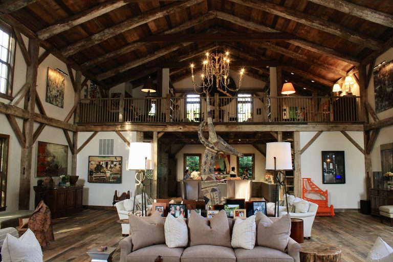 The great room of a timber-framed Hamptons home, featuring soaring vaulted ceilings, an upper balcony, a wrought-iron chandelier, and an eclectic collection of contemporary art and sculpture.