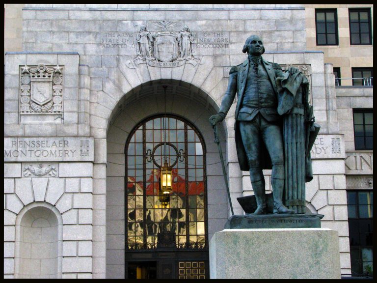 A bronze statue of George Washington stands before the New York State Capitol, beneath the state's Excelsior coat of arms.